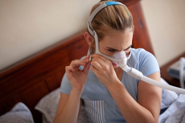Woman adjusting CPAP mask in bed, illustrating sleep apnea treatment and its impact on oral health.