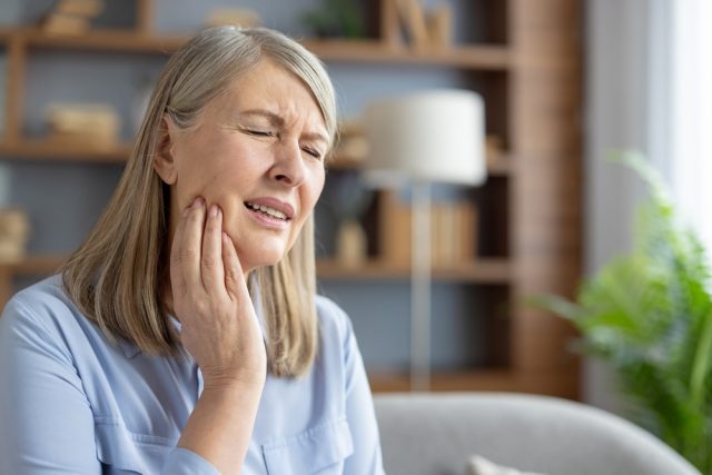 Woman experiencing jaw pain, holding her face in discomfort, indicating symptoms of TMJ disorder, with a cozy indoor setting in the background.